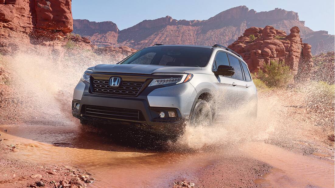 Passenger-side front 3/4 view of 2019 Honda Passport Elite, in Deep Scarlet Pearl, driving on rugged mountain road with accessory roof basket and running boards. 
