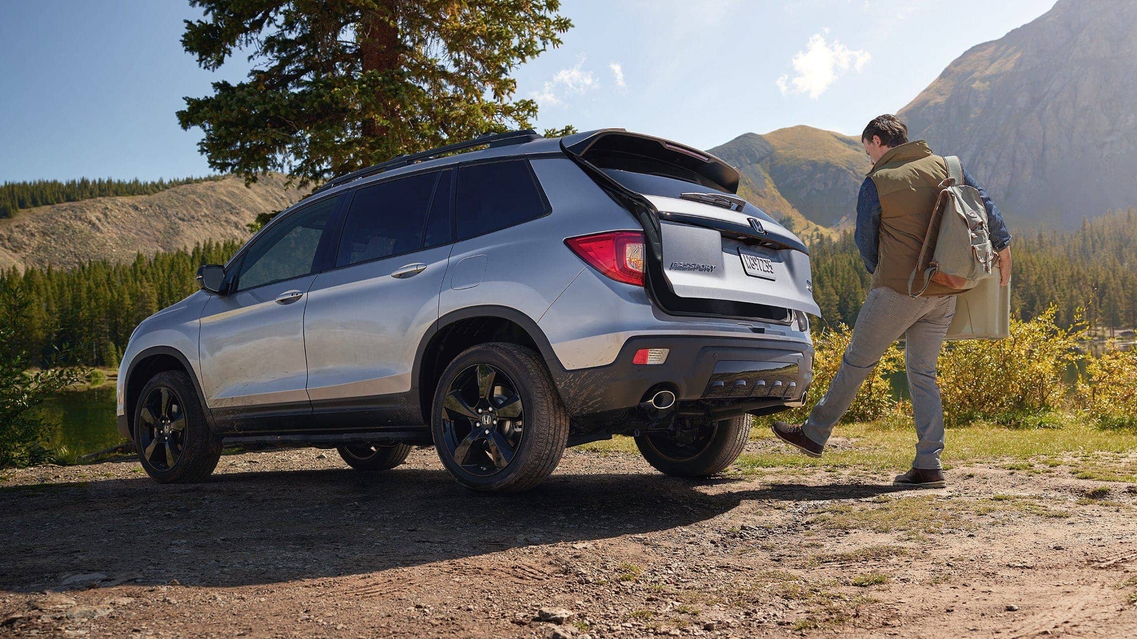Driver-side rear 3/4 view of 2019 Honda Passport Elite in Lunar Silver Metallic, parked at campsite and demonstrating the hands-free access power tailgate feature.