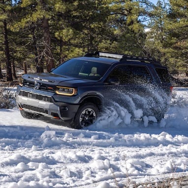 2027 Honda Passport TrailSport Elite in Smoke Blue Pearl front driving through deep snow in a pine forest (Mid-Size SUV).