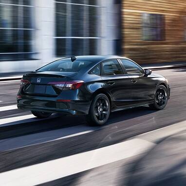 2027 Honda Civic Sedan Sport Hybrid in Crystal Black Pearl rear three-quarter view driving on a city street (Compact Sedan)